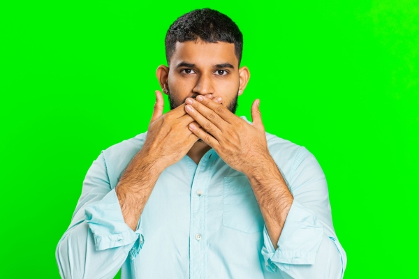 An Asian man with short dark hair hold his hands up to his mouth as if silencing himself. He wears a light blue shirt. The background is green